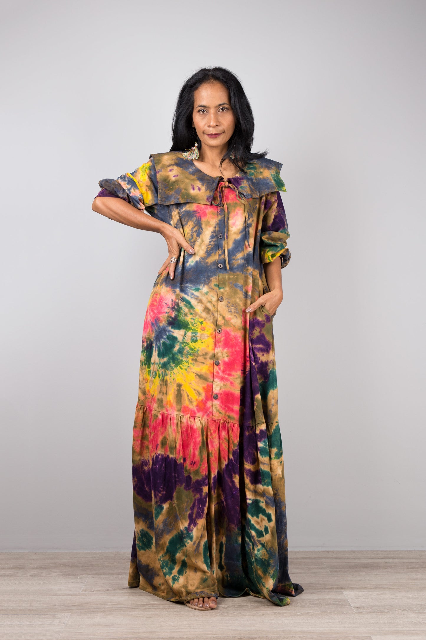 Woman in a vibrant tie-dye kaftan maxi dress with navy collar and billowy sleeves, posed against a gray wall and wooden floor, styled with nude sandals and dangling earrings


