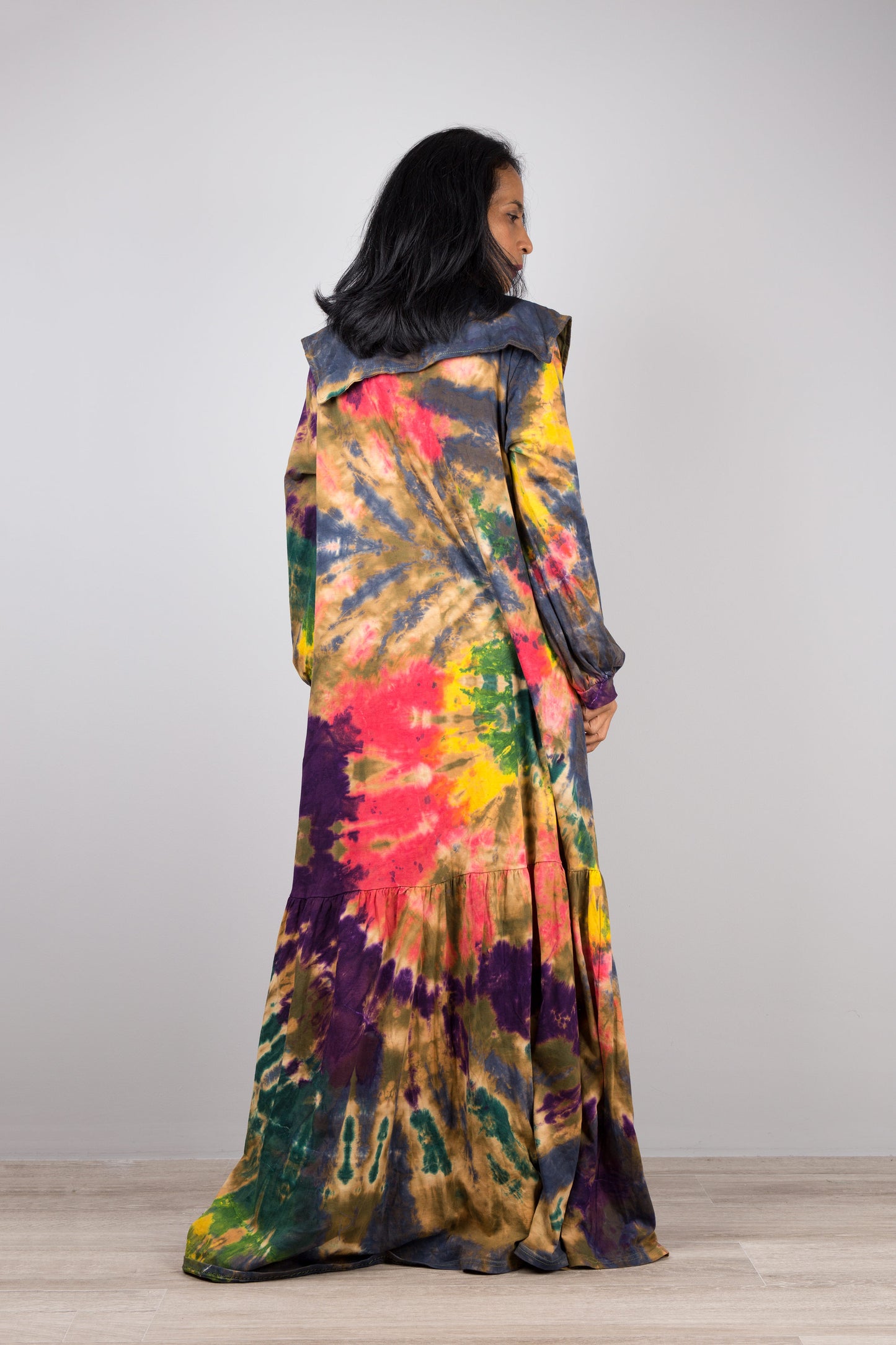 Tie-dye kaftan maxi dress with a navy-trimmed V-neck and long sleeves, worn by a woman with nude sandals and earrings, set against a minimalist gray and wood interior.

