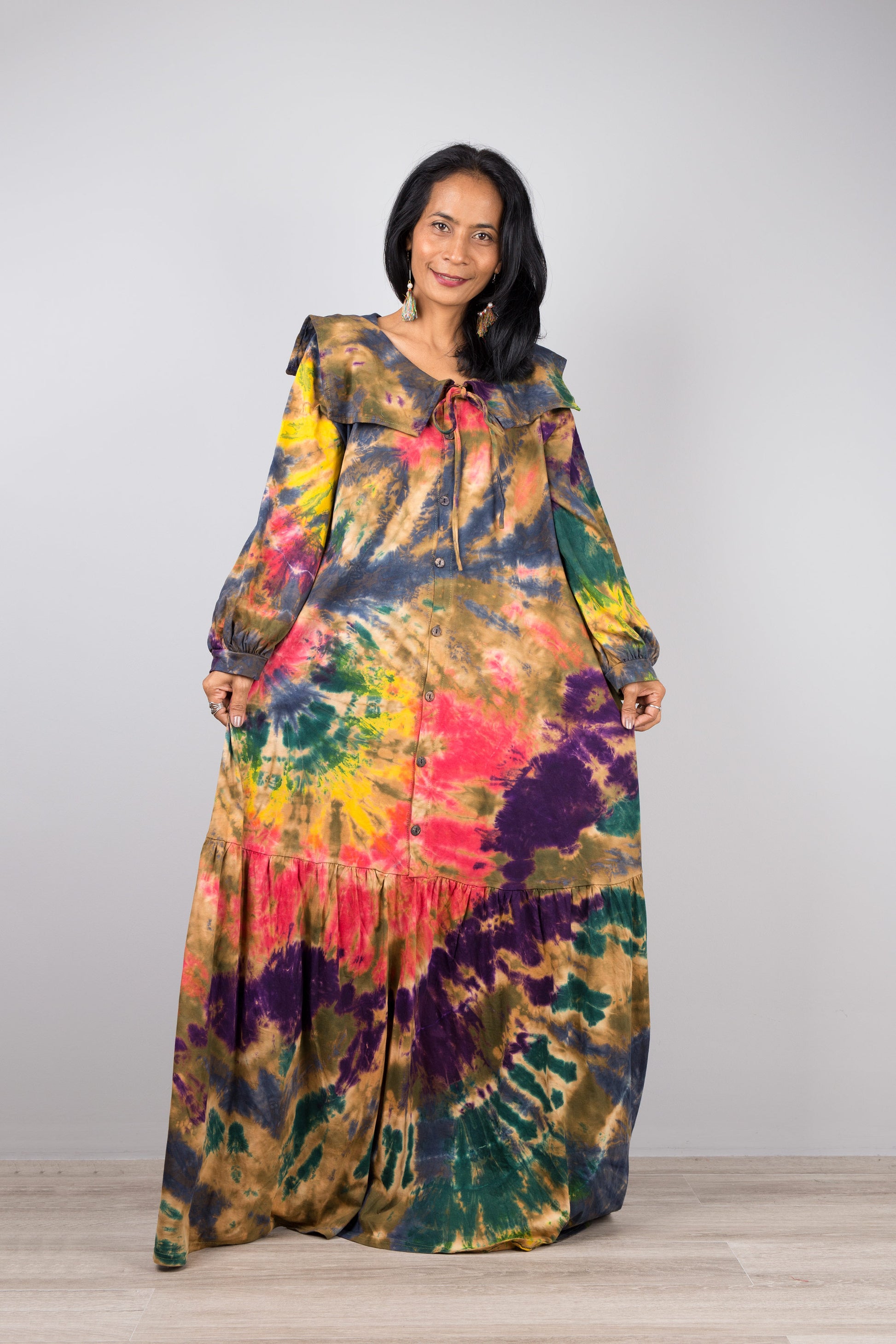 A woman wearing a tie-dye kaftan maxi dress with navy trim, V-neck, and long sleeves, standing against a gray wall with a wooden floor, accessorized with nude sandals and dangling earrings.

