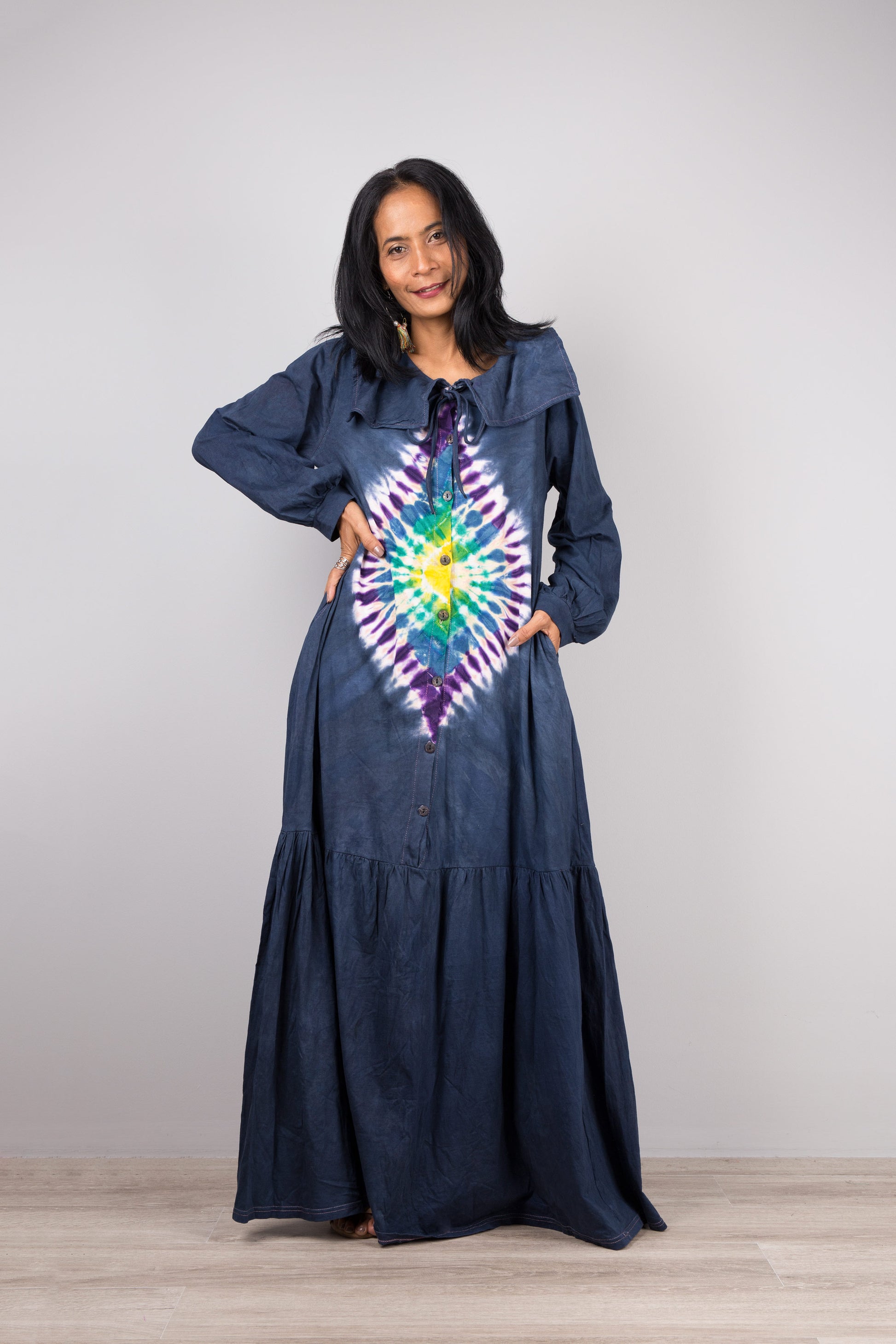 A woman wearing a dark blue tie-dye kaftan maxi dress with navy trim, deep V-neck, and long sleeves, standing against a gray wall with a wooden floor, accessorized with nude sandals and dangling earrings

