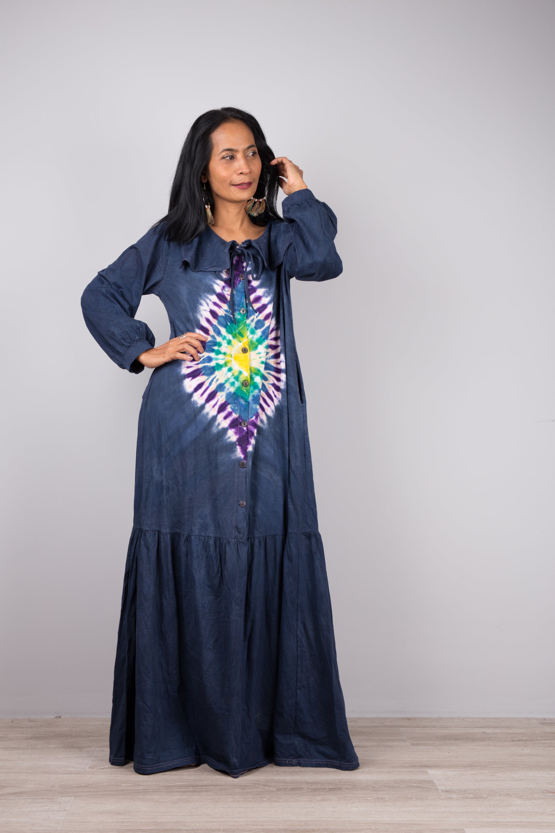 Stylish woman in a floor-length dark blue tie-dye kaftan dress with navy trim, V-neck, and billowy sleeves, posed against a gray wall and wooden floor, wearing nude sandals and earrings

