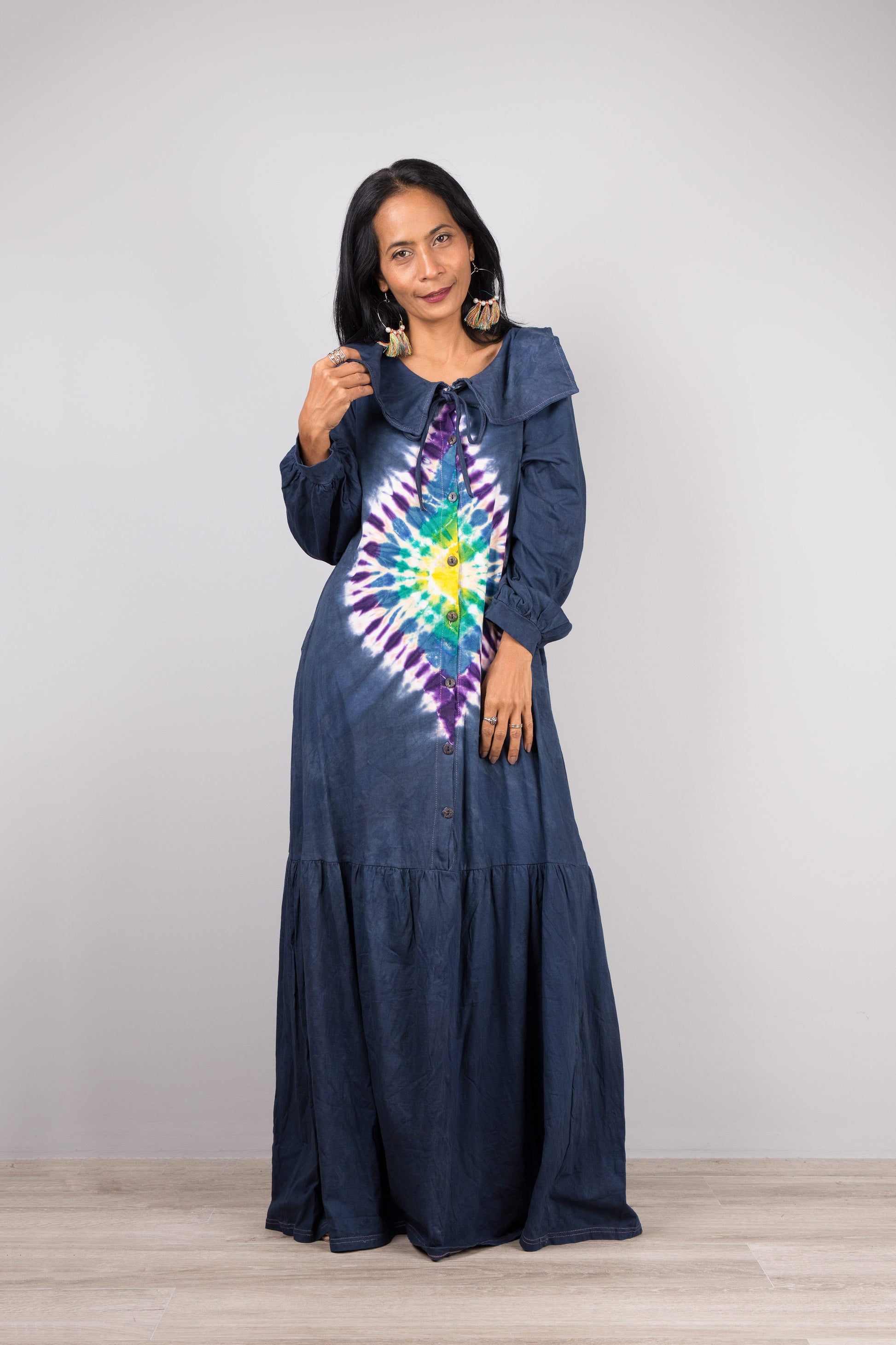 Nuichan dark blue tie-dye kaftan maxi dress with a navy-trimmed collar, deep V-neck, and long sleeves, modeled by a woman with nude sandals and dangling earrings, against a simple gray and wood backdrop


