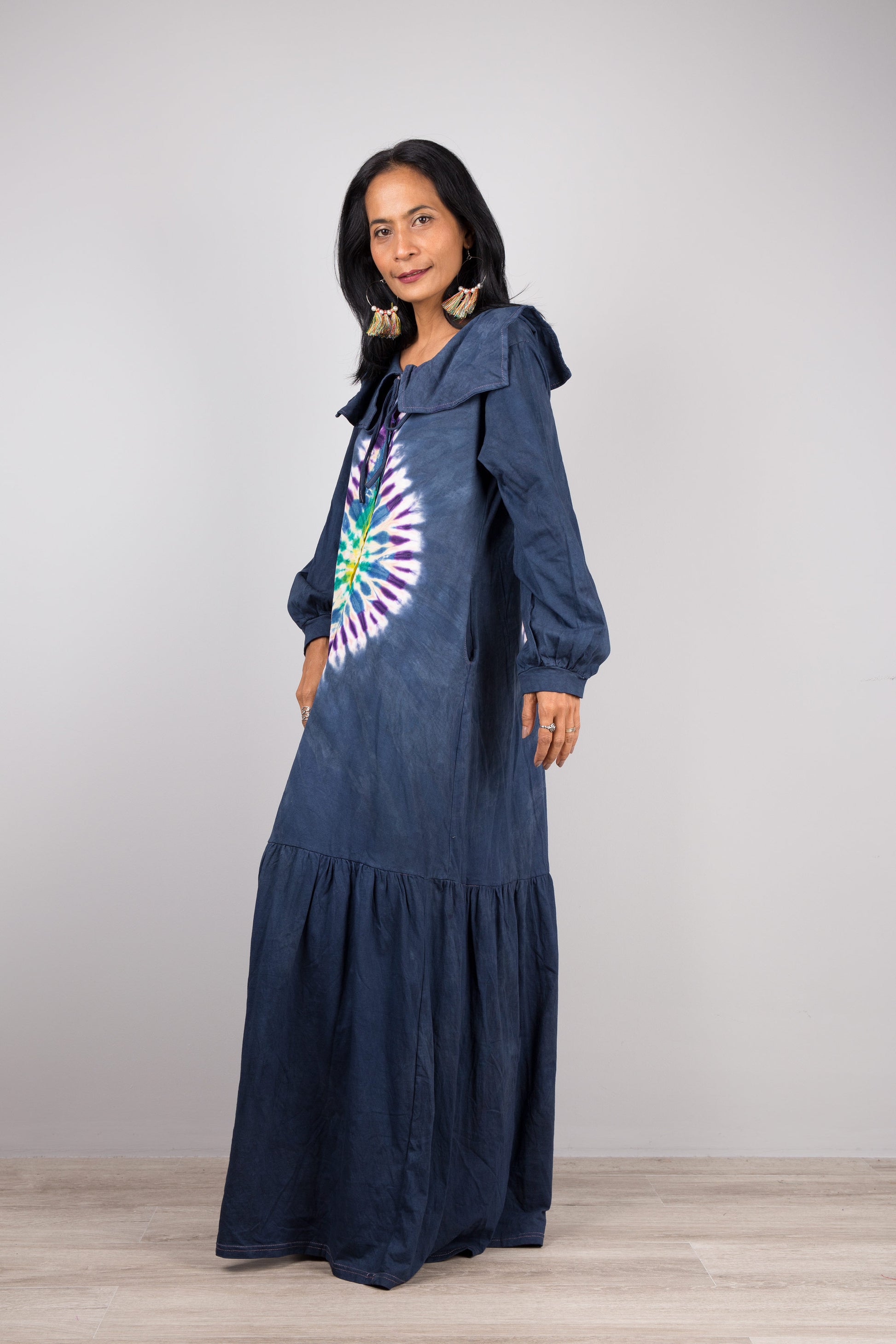 Woman showcasing a dark blue tie-dye kaftan maxi dress with navy trim, flowing design, deep V-neck, and long sleeves, accessorized with nude sandals, on a gray and wooden floor setting

