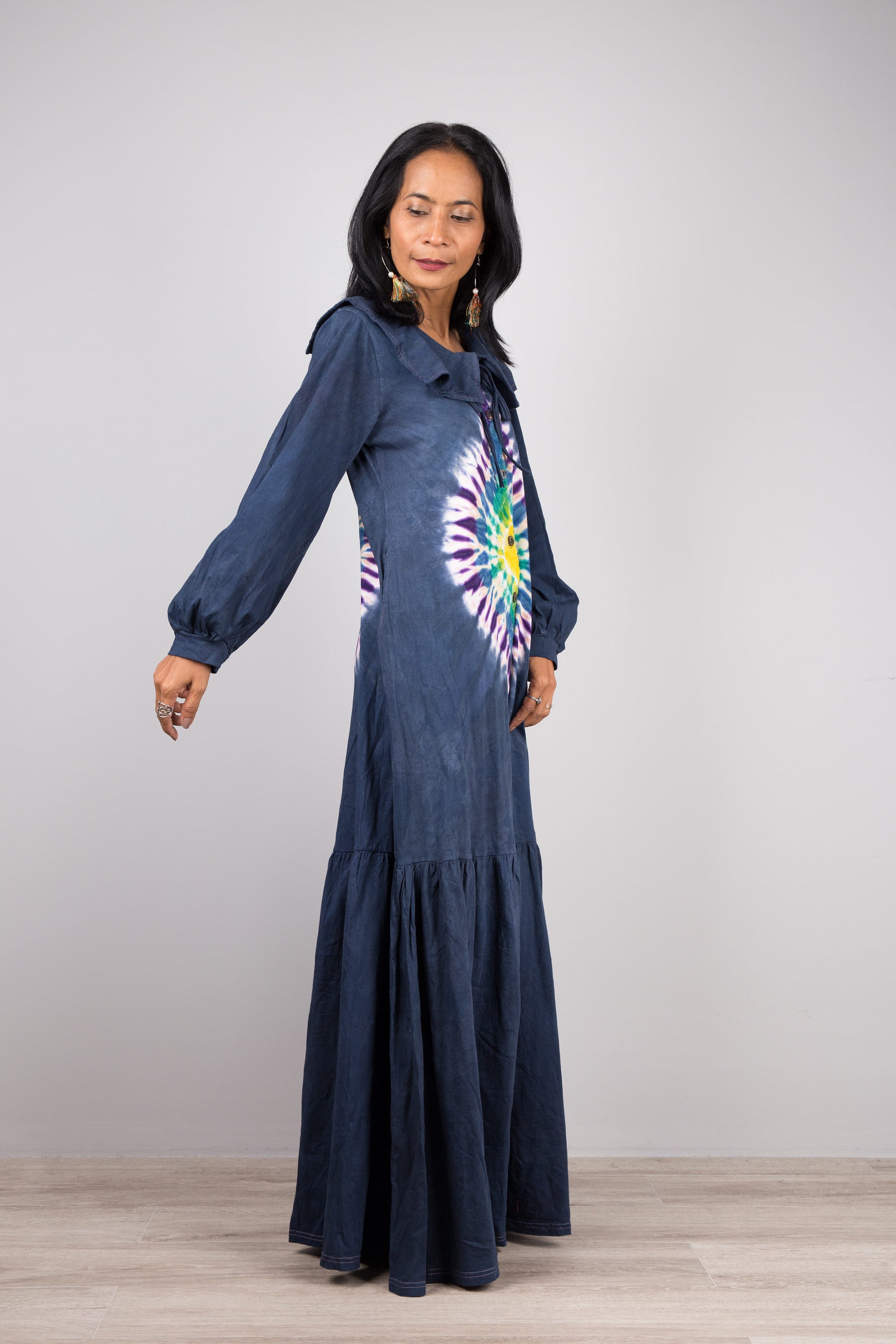 Elegant dark blue tie-dye kaftan maxi dress with a navy-trimmed collar and gathered sleeves, worn by a woman against a gray wall and wooden floor, paired with nude sandals and earrings.

