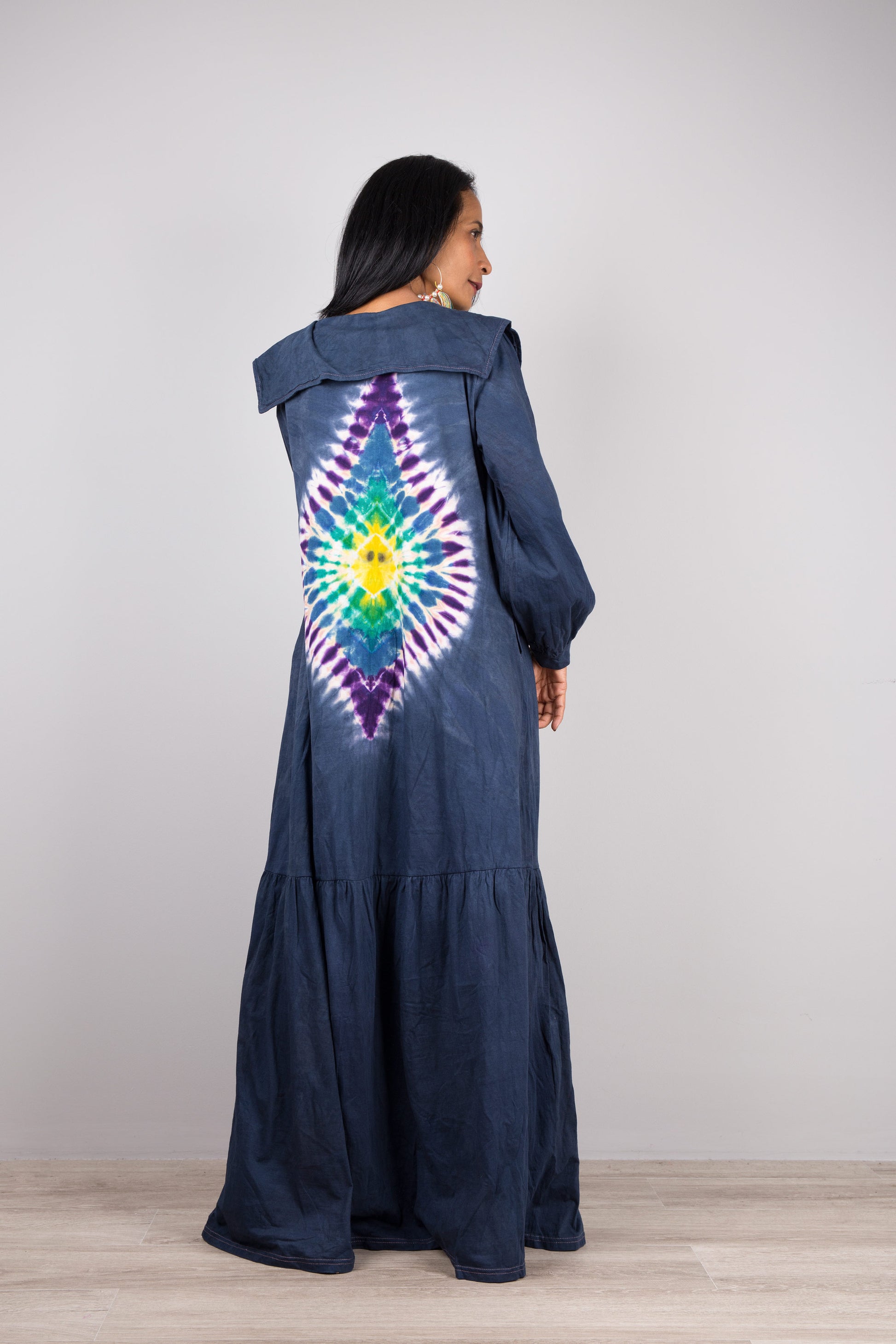 Model in a dark blue tie-dye pattern kaftan maxi dress with navy trim, deep V-neck, and long sleeves, standing on a wooden floor with a gray wall background, wearing nude sandals

