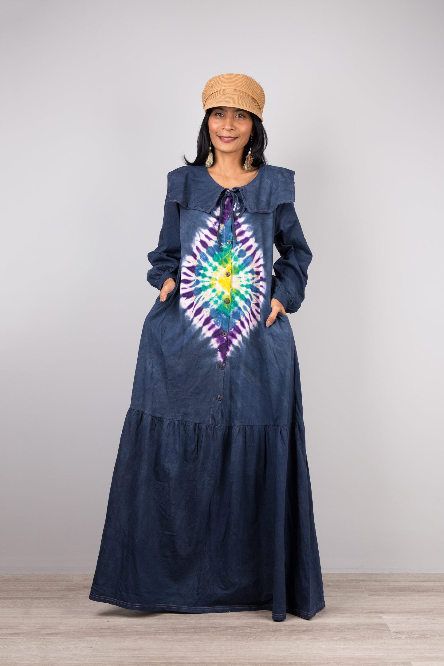 Woman in a vibrant dark blue tie-dye kaftan maxi dress with navy collar and billowy sleeves, posed against a gray wall and wooden floor, styled with nude sandals and dangling earrings

